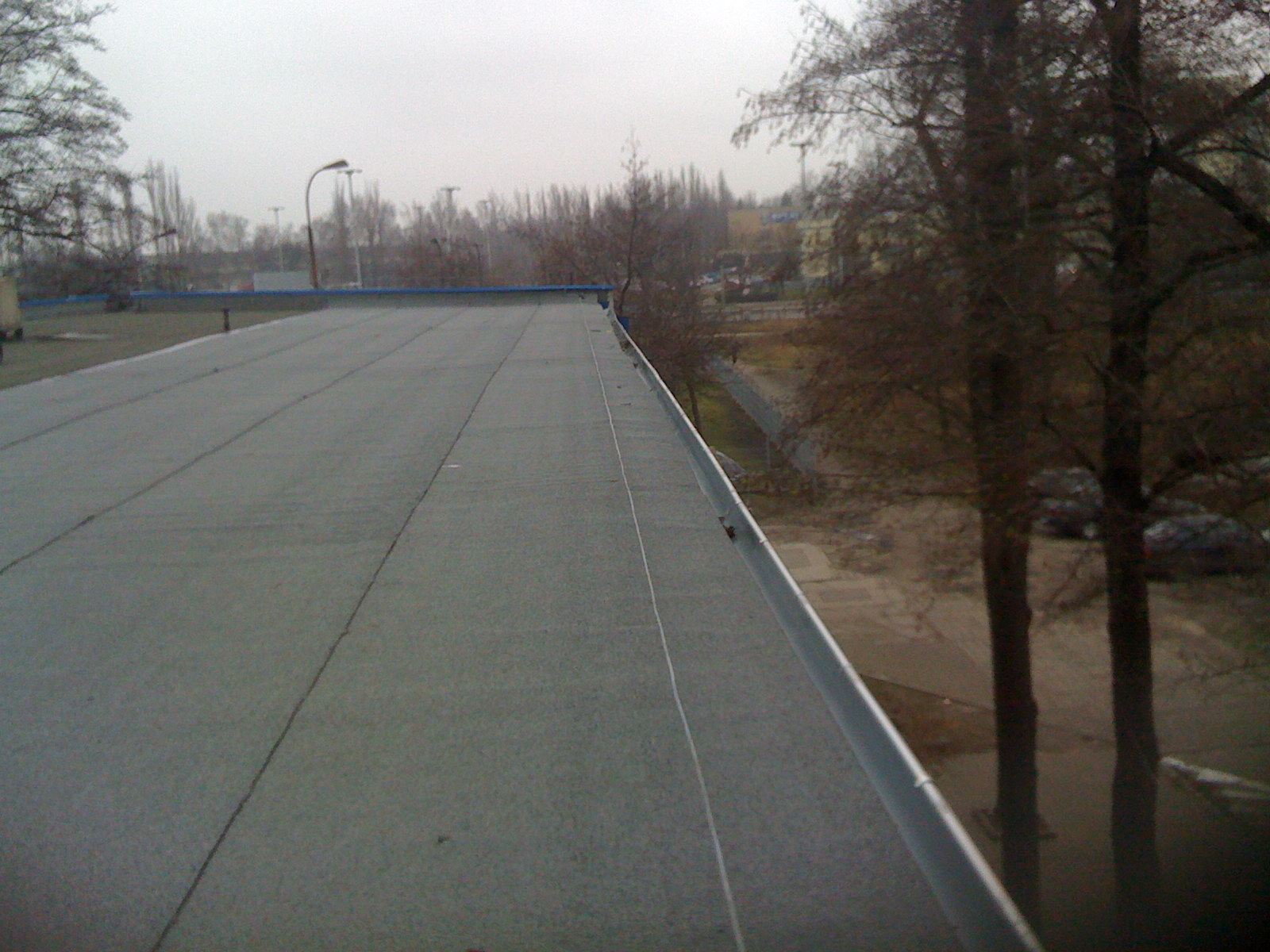 Szary, płaski dach budynku w Chorzowie z widoczną metalową obróbką blacharską, w tle drzewa i zabudowa miejska w pochmurny dzień.